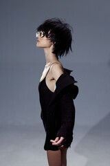 Stylish woman in black dress wearing sunglasses with short hair posing on gray background