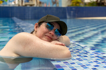 A serene woman wearing a black baseball cap and sunglasses relaxes in a turquoise pool. This image...