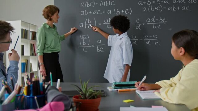 Teacher and African American student pointing to mathematical equation on chalkboard filled with algebraic expressions, as other students attentively participating in math lesson