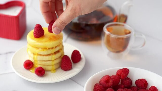 Hand placing fresh raspberry on top of mini pancake stack with honey drizzle and berries served with tea on white table for breakfast. Homemade gourmet breakfast, food styling, culinary presentation.