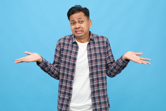 Sad young asian man wearing casual clothes, spreading his arms, shrugging his shoulders, looking confused, clueless, isolated on blue background.