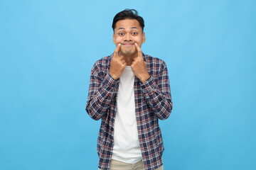 Young Asian man wearing casual clothes, showing a sweet smile with index finger on the tip of his...