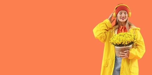 Young woman in raincoat holding autumn flowers and listening to music on orange background with...