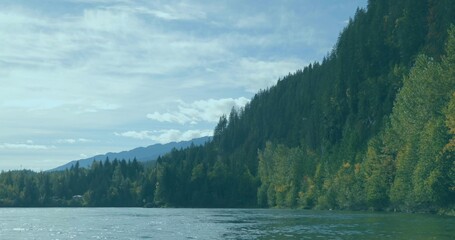 Flowing river reflecting evergreen hillside and distant mountains in mountain park under cloudy sky