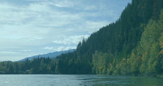 Fototapeta Flowing river reflecting evergreen hillside and distant mountains in mountain park under cloudy sky