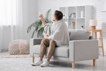 Handsome young man with air conditioner remote control sitting on sofa in living room