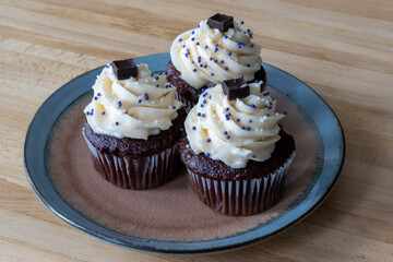 Three chocolate cupcakes with vanilla icing, sprinkles, and a chocolate square on a small plate and wooden table. The homemade baked and frosted brownie style cakes swirl in buttercream icing. 