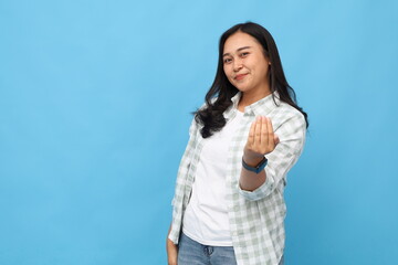 Fototapeta premium Young asian woman wearing casual clothes, inviting or calling to come or join with hand, friendly facial expression smiling happily looking at camera, isolated blue background.