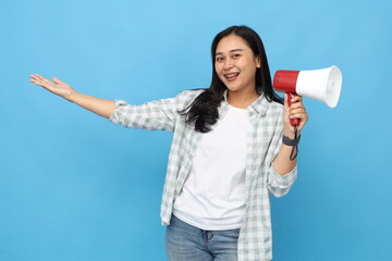 Beautiful and cheerful Asian woman speaking into a megaphone while presenting something sideways towards empty space, standing isolated on blue background.