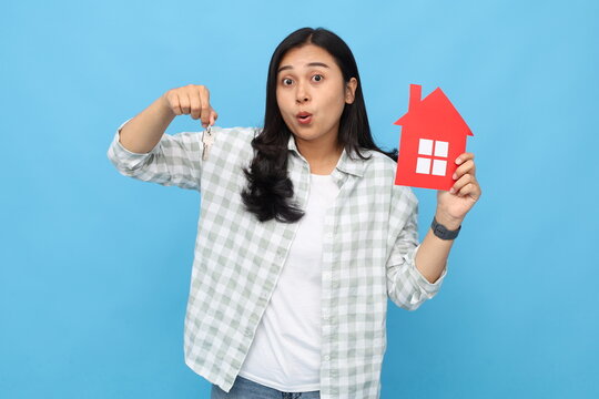 Female hands holding a small red paper house image and house keys, posing on a blue background wall in a studio with copy space for promotional or design content, real estate and family home concept.