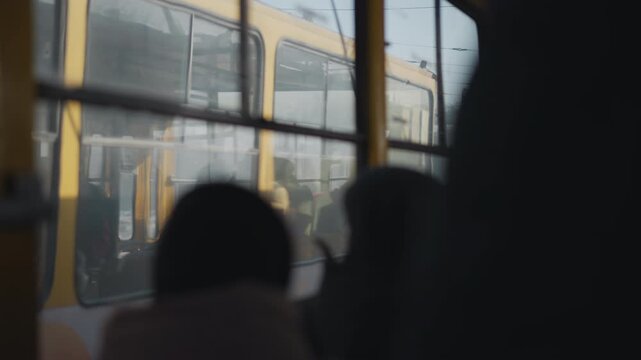 Inside moving bus, passengers in winter coats watch opposite tram glide past outside, view through windows with soft blur on glass, muted city light, motion on snowy street during urban commute