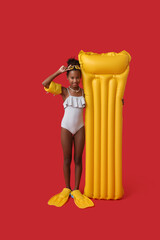Obraz premium Cute little African-American girl in swimsuit with flippers and inflatable mattress on red background
