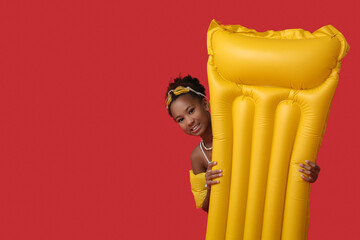 Cute little African-American girl in swimsuit with inflatable mattress on red background