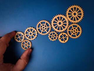 The image shows a hand placing a wooden gear, which is part of a larger arrangement of gears on a blue background