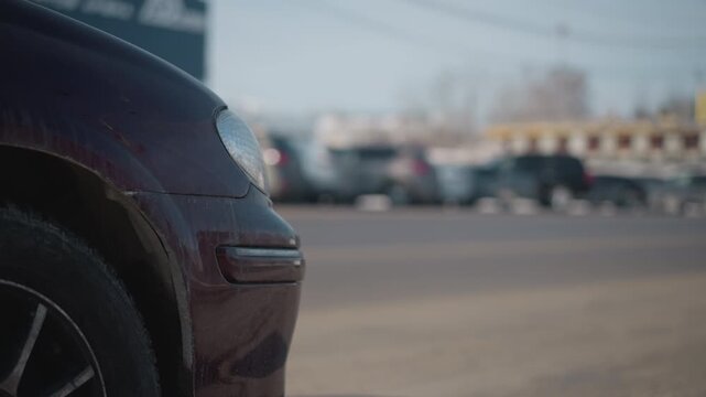 close up maroon car headlight and bumper showing flecks of mud parked on busy urban street with moving cars driving by under bright winter daylight, asphalt pavement and blurred city background