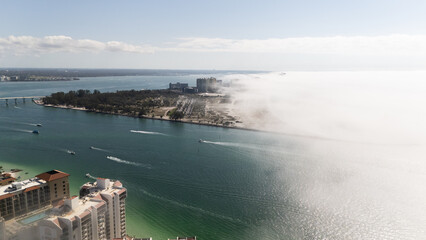Aerial view of coastal city with boats on turquoise water and thick fog rolling over shoreline, symbolizing nature, mystery, travel, weather phenomena, and scenic urban waterfront landscape.