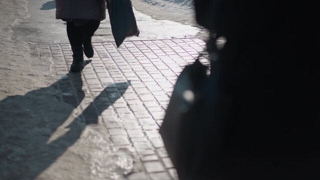 leg level view shows people in sturdy boots walking along icy pavement, long shadows stretching over wet bricks, winter light glinting off slush and snow, city traffic moving past curb in morning.