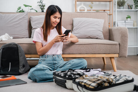 A young woman sits on the floor packing a suitcase while using her smartphone, preparing for a trip in a cozy living room.