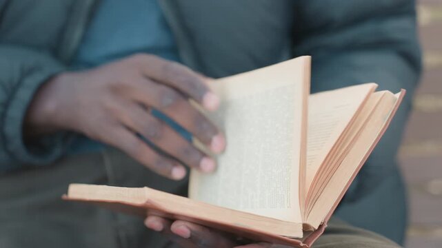 Close up of adult reading novel outdoors, hands holding worn book on lap, pages turning to new chapter under soft daylight with blurred background