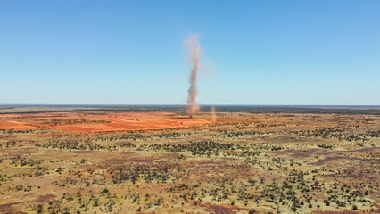 This aerial photograph captures a dust devil swirling across a vast, arid landscape. The clear blue...