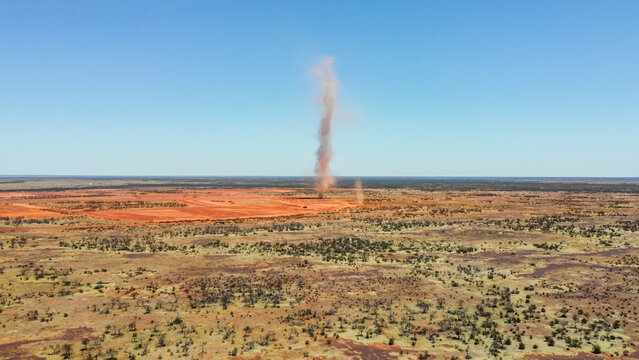 This aerial photograph captures a dust devil swirling across a vast, arid landscape. The clear blue sky provides a stark contrast to the red earth and sparse vegetation, with the swirling column.
