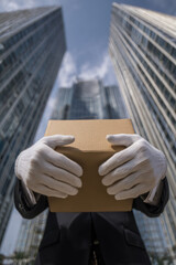 A white-gloved hand raises a cardboard box against a backdrop of modern skyscrapers, symbolizing delivery, service, and logistics in the city.
