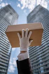 A white-gloved hand raises a cardboard box against a backdrop of modern skyscrapers, symbolizing delivery, service, and logistics in the city.
