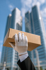 A white-gloved hand raises a cardboard box against a backdrop of modern skyscrapers, symbolizing delivery, service, and logistics in the city.
