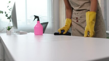 Professional cleaner wearing yellow protective gloves and beige apron sanitizing white desk surface using spray bottle and cleaning rag in office workplace. - Powered by Adobe
