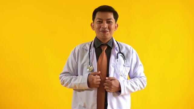 Young male doctor straightening his clothes and waving his hand forward isolated on yellow background