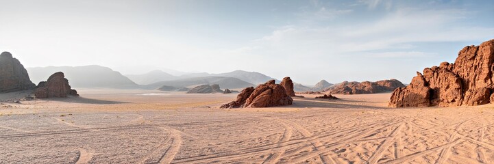 dramatic dramatic mountainous desert landscape, vast sandy desert with rugged red rock formations, cloudy blue sky in the background, vehicle tracks in the foreground, wide panoramic view
