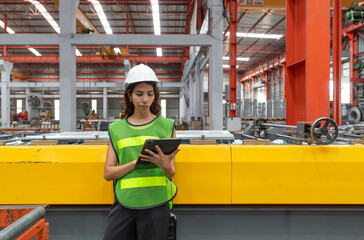 A young woman wearing a safety vest and helmet stand in a busy warehouse, manage online document on...