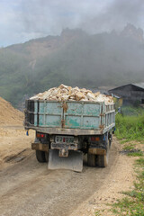 A heavy-duty dump truck, loaded with rocks, is parked on a dirt road at a mining site with a hazy, mountainous background.