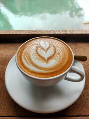 A beautifully crafted latte art in a white cup, resting on a wooden surface, with a serene green background reflecting water.