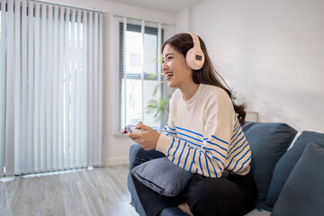 Obraz premium A young woman sits on a sofa wearing headphones while holding a game controller and playing a video game on her laptop. Her cheerful expression shows enjoyment and excitement.