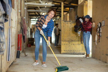 Female employee cleans stable, sweeps floors with broom, and carries out cleaning activities. She...