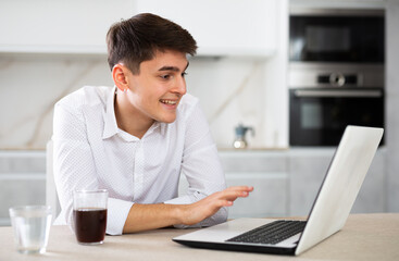 Smiling young man making video call on laptop at table at living room, chatting with relatives or...