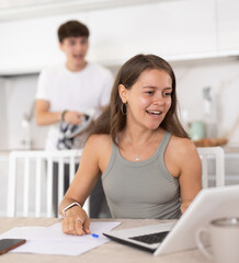 Smiling young lady doing business on notebook and speaking to her husband standing at the kitchen...