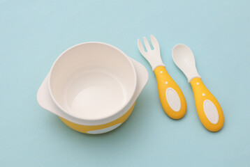 Bowl with spoon and fork on blue pastel background. Accessories for feeding small children. Top view