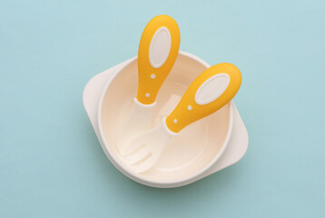 Bowl with spoon and fork on blue pastel background. Accessories for feeding small children. Top view