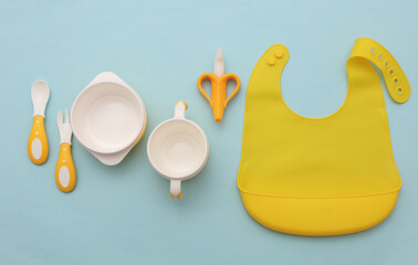 Silicone baby bib with bowls and spoons on blue pastel background. Accessories for feeding a small children. Top view