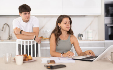 Fototapeta premium Annoyed young girl doing business on notebook while guy is brawling with her at the kitchen table