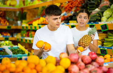 Young positive couple choosing fresh pineapples and oranges tropical fruits in supermarket
