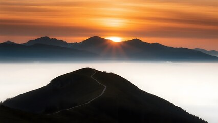 Scenic mountain range silhouette at sunrise with sun rays breaking through the clouds over a misty valley and a winding path on a foreground hill