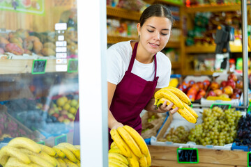Smiling female merchandiser in uniform laying out bananas at grocery supermarket