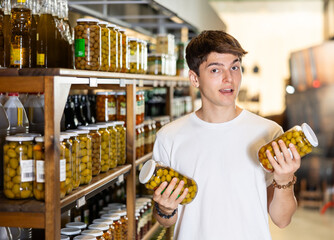 Interested young guy standing in supermarket aisle next to shelves filled with pickled olives,...