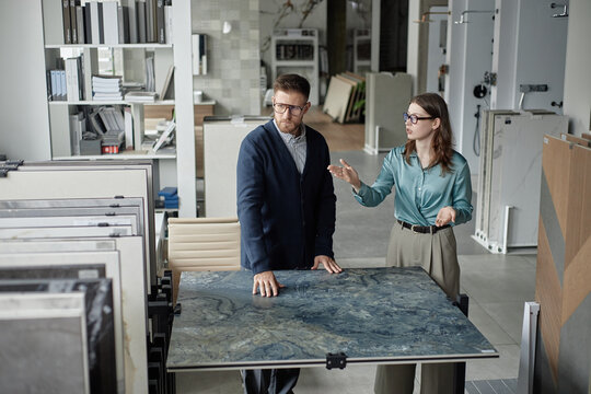 Caucasian man and woman discussing large stone countertop sample in modern showroom, man examining surface while woman gesturing and explaining options