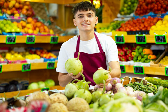 Smiling young seller man in apron demonstrating small green pumpkin in glocery shop