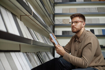 Caucasian man wearing glasses selecting ceramic tiles from display shelves, holding two samples in hands, examining surface texture and comparing options in store