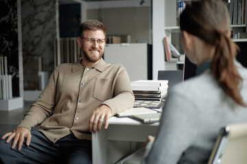Caucasian young adult man smiling and talking with Caucasian young adult woman in modern office setting, both sitting at desk with stack of books and open notebook visible